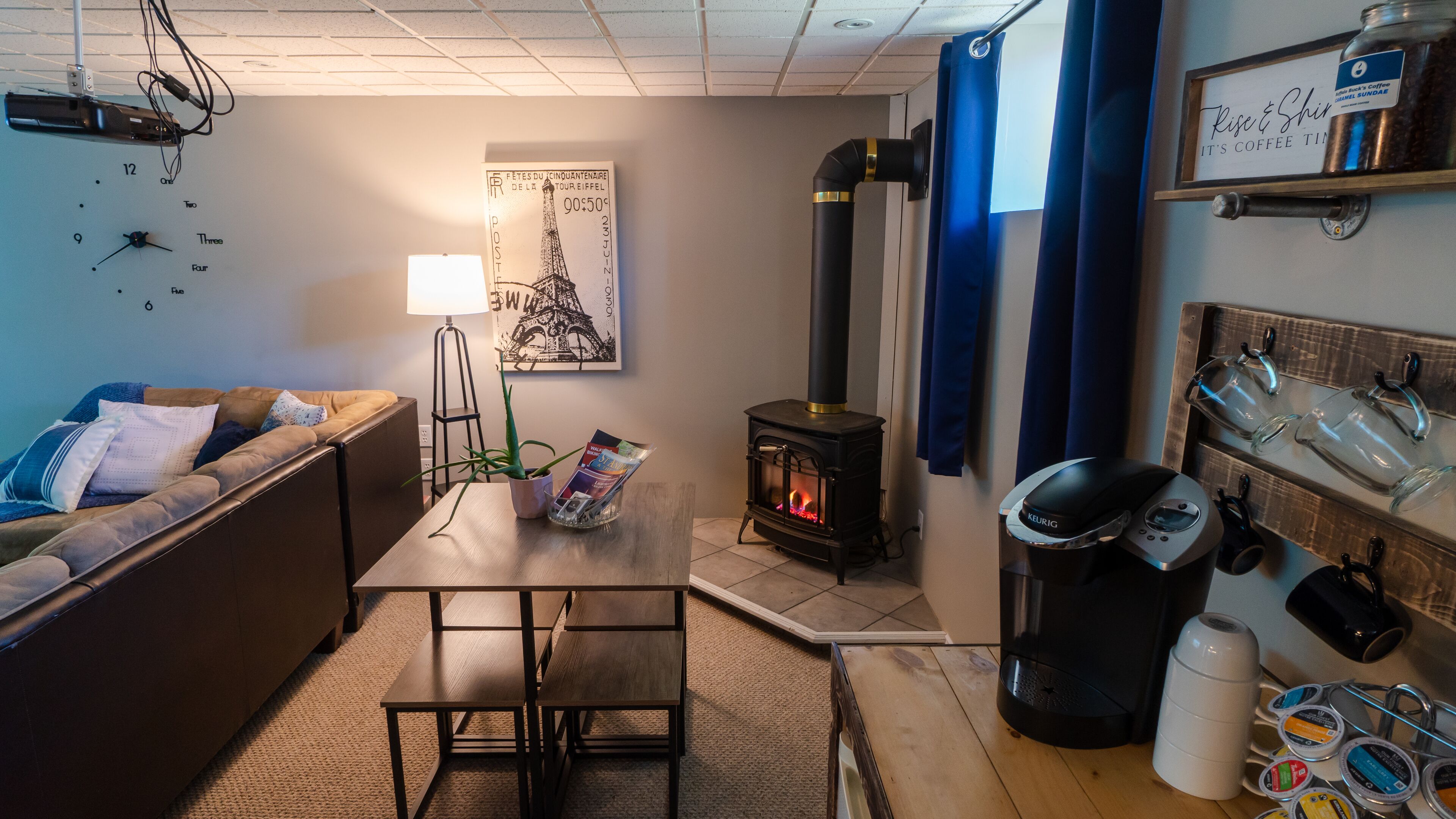 A cozy living room with a sofa, a coffee table, a small table with magazines, a wall-mounted lamp, a poster of the Eiffel Tower, a wood-burning stove, and a coffee machine.