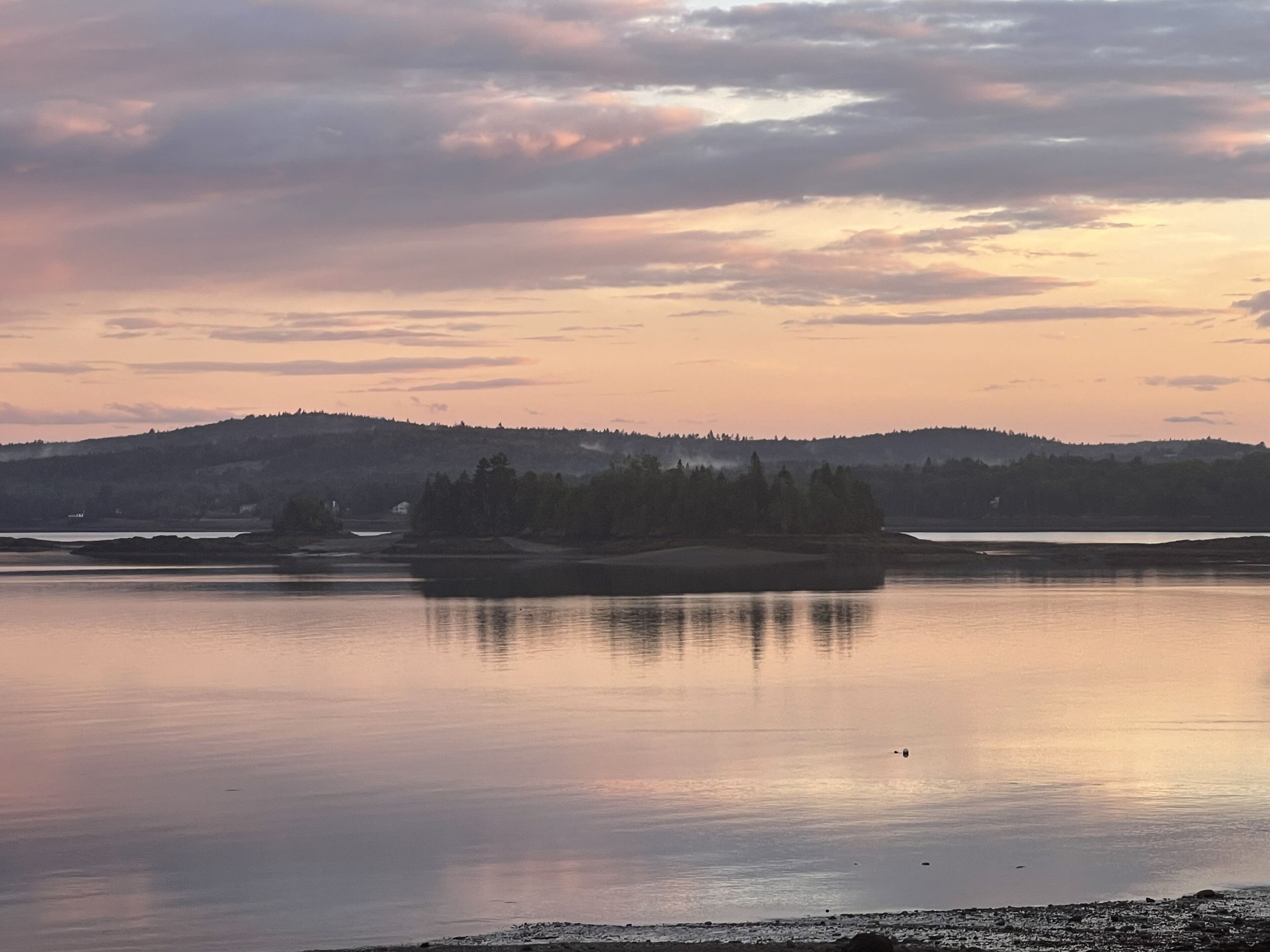 Ein ruhiger See mit einer kleinen Insel in der Mitte, umgeben von Hügeln und einem bewölkten Himmel.