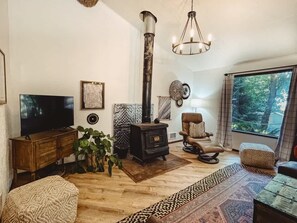 A cozy living room with a large wooden TV stand, a black wood-burning stove, a comfortable leather armchair, and a large window with a view of trees. - Forested Cottage in the Woods, near Olympic Park (Port Angeles)