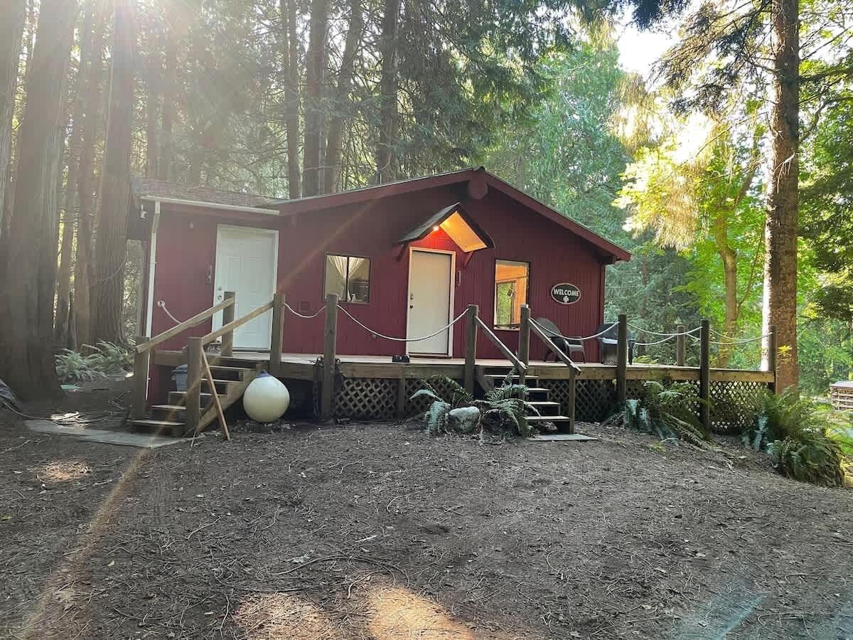 A small, rustic cabin with a wooden deck and a welcome sign, surrounded by dense forest.
