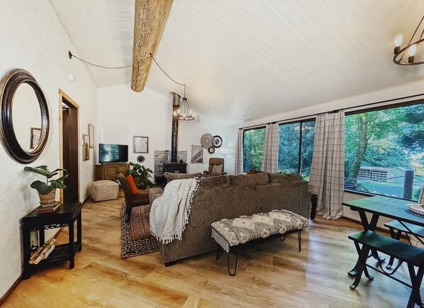 A cozy living room with a large sofa, a coffee table, and a dining table. There is a television, a fireplace, and a large window with curtains.