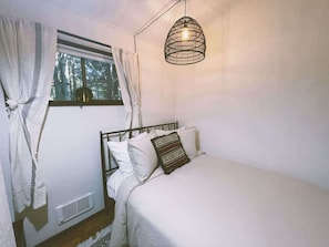 Un dormitorio con cama, cortinas blancas, una ventana con vista a árboles y una lámpara colgante.