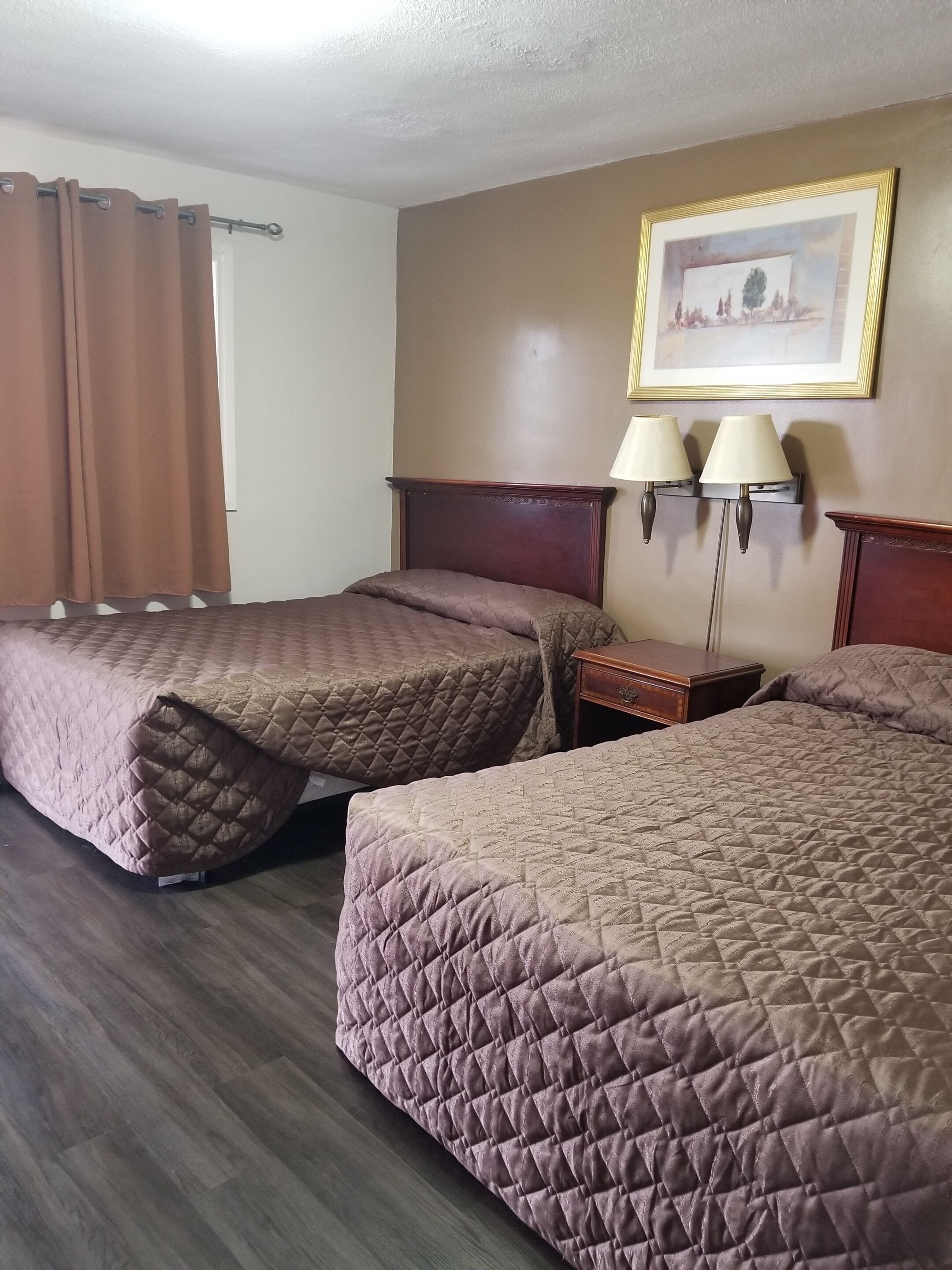 A hotel room with two queen beds, a wooden headboard, a nightstand, two wall-mounted lamps, and a framed picture on the wall.