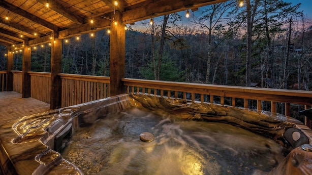 Ein Whirlpool mit Holzterrasse und Lichterkette, umgeben von bewaldeter Landschaft.