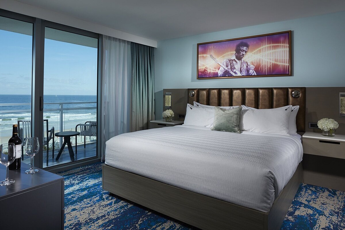 Une chambre d’hôtel avec un grand lit, une vue sur l’océan, un coin repas avec une table et des chaises, et un tableau accroché au mur.