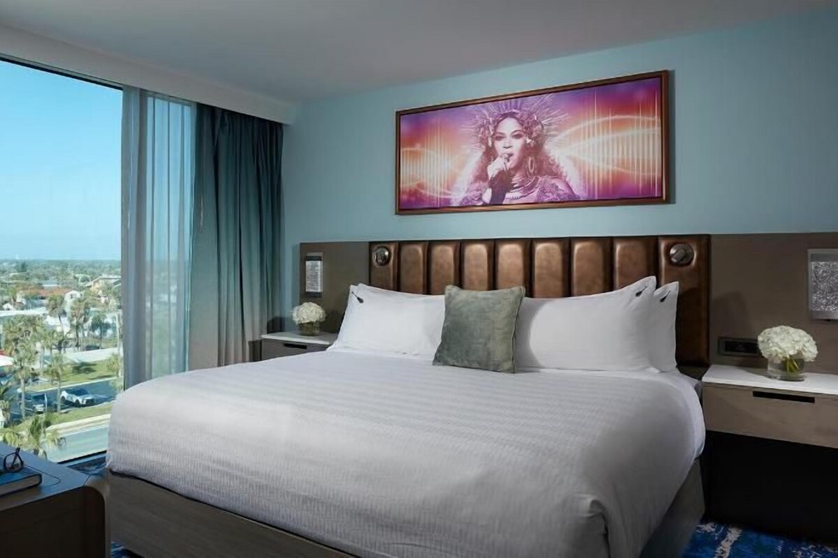 Une chambre d’hôtel avec un grand lit, un téléviseur sur le côté gauche, une fenêtre donnant sur la ville et un tableau au-dessus du lit.