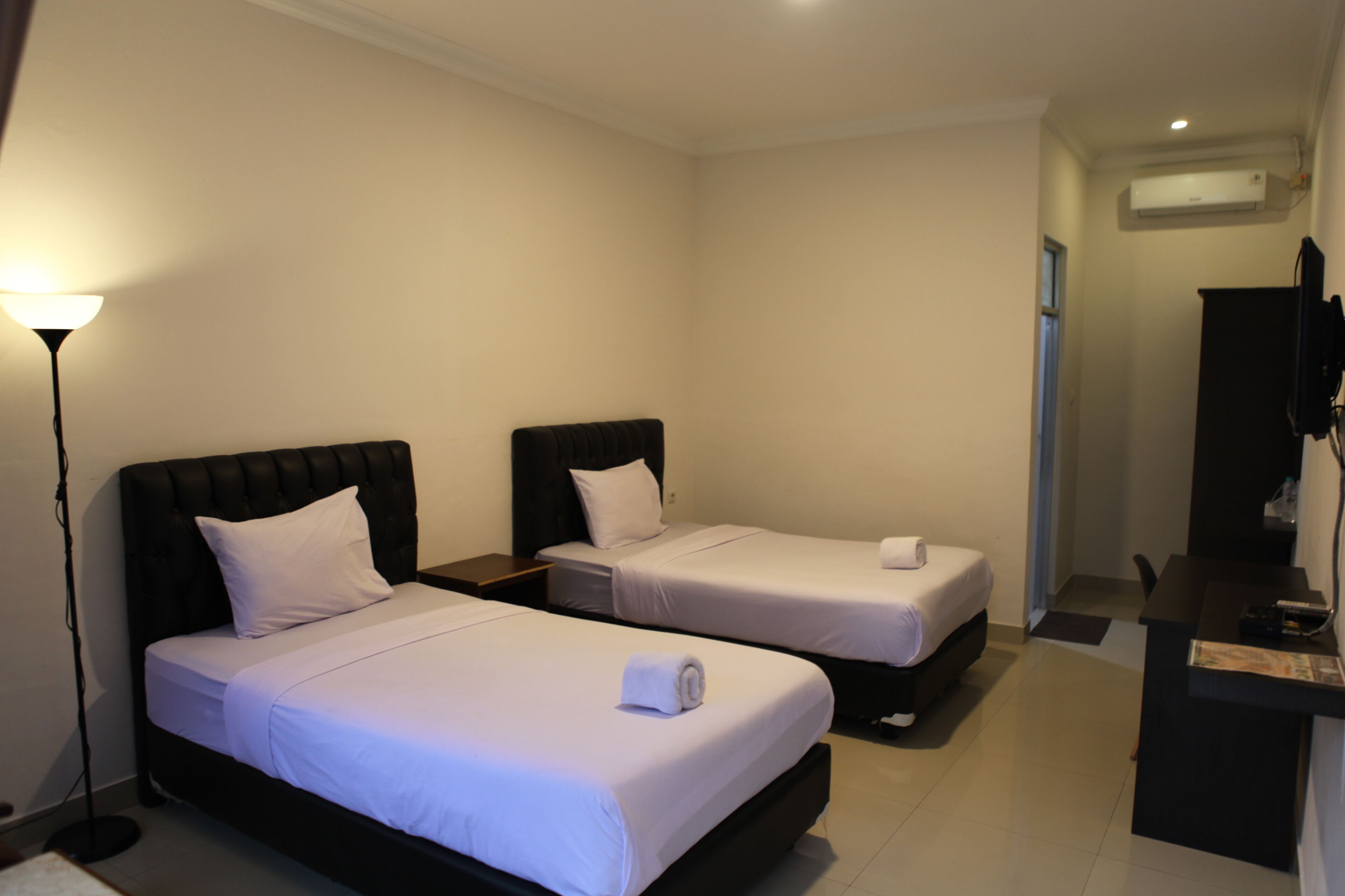 A hotel room with two beds, a desk, a chair, a lamp, and an air conditioning unit.