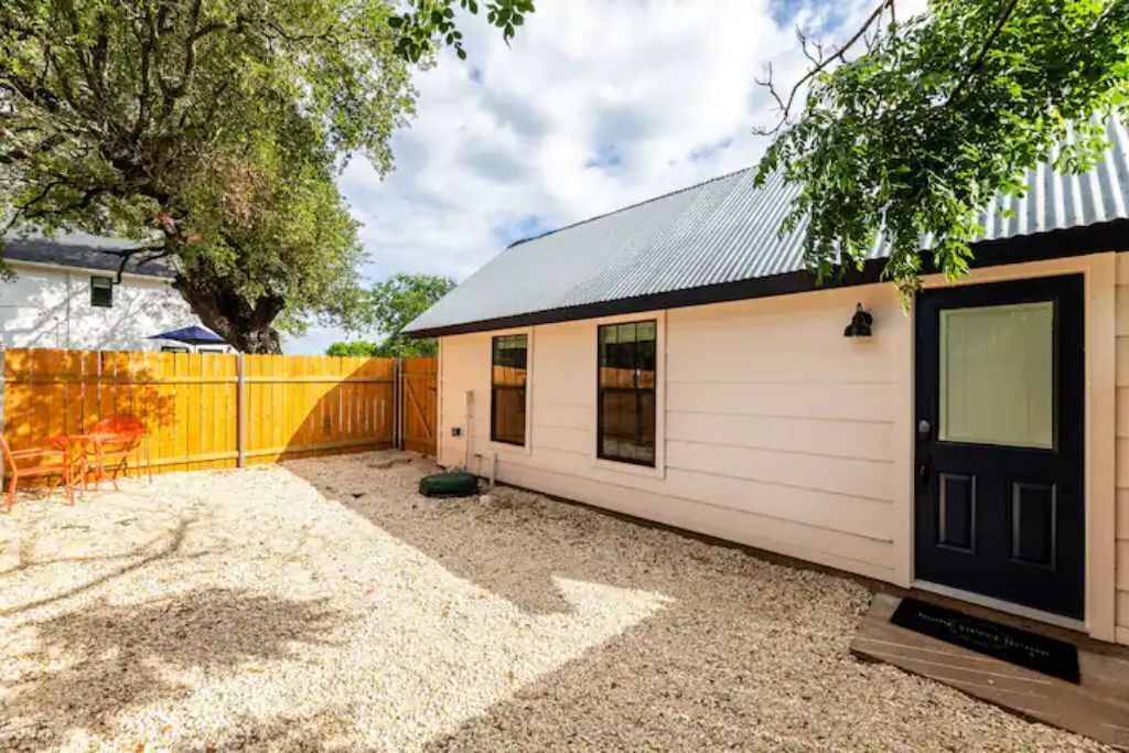 A single-story building with a gravel driveway, a wooden fence, and a large tree providing shade.