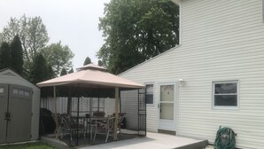 Un patio couvert avec une table et des chaises, un abri de jardin et une maison avec un garage et une fenêtre.
