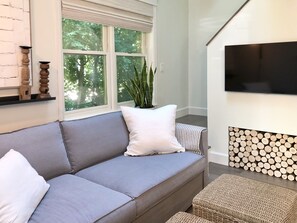 A living room with a grey sofa, white cushions, a potted plant, a flat-screen TV mounted on the wall, and a stack of logs against the wall.