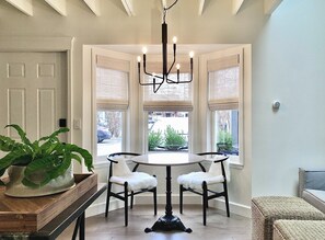 A dining area with a round table and four chairs, a chandelier, and a view through a bay window.