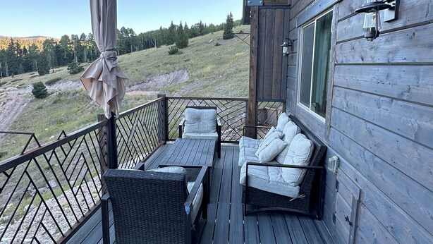 Una terraza de madera con mobiliario exterior, vistas a un paisaje montañoso y un edificio de madera con ventana y un aplique de pared.