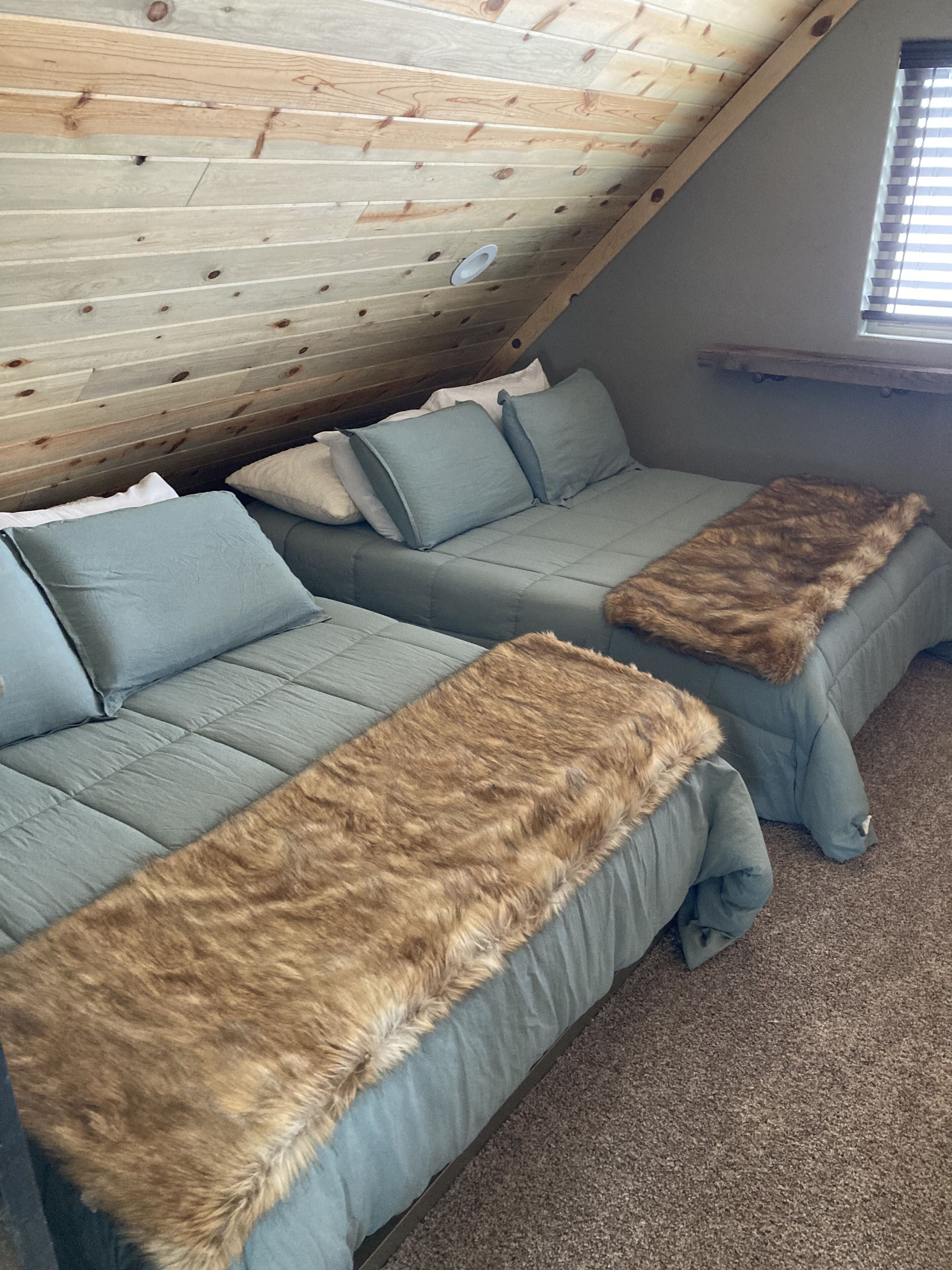 Ein gemütlicher Raum mit schrägen Wänden und Holzvertäfelung. Zwei graue Sofas mit Felldecken und ein kleines Fenster mit Jalousien sind zu sehen.