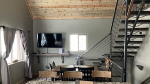 Una habitación con una mesa de mármol, sillas, un televisor, una escalera y una ventana con cortinas.