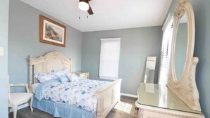 Une chambre à coucher comprenant un lit, une commode, une chaise, un ventilateur de plafond, une fenêtre avec des stores et un tableau encadré accroché au mur.