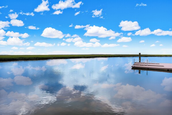 A calm lake with a dock extending into the water, surrounded by a flat landscape and a clear blue sky with scattered clouds.