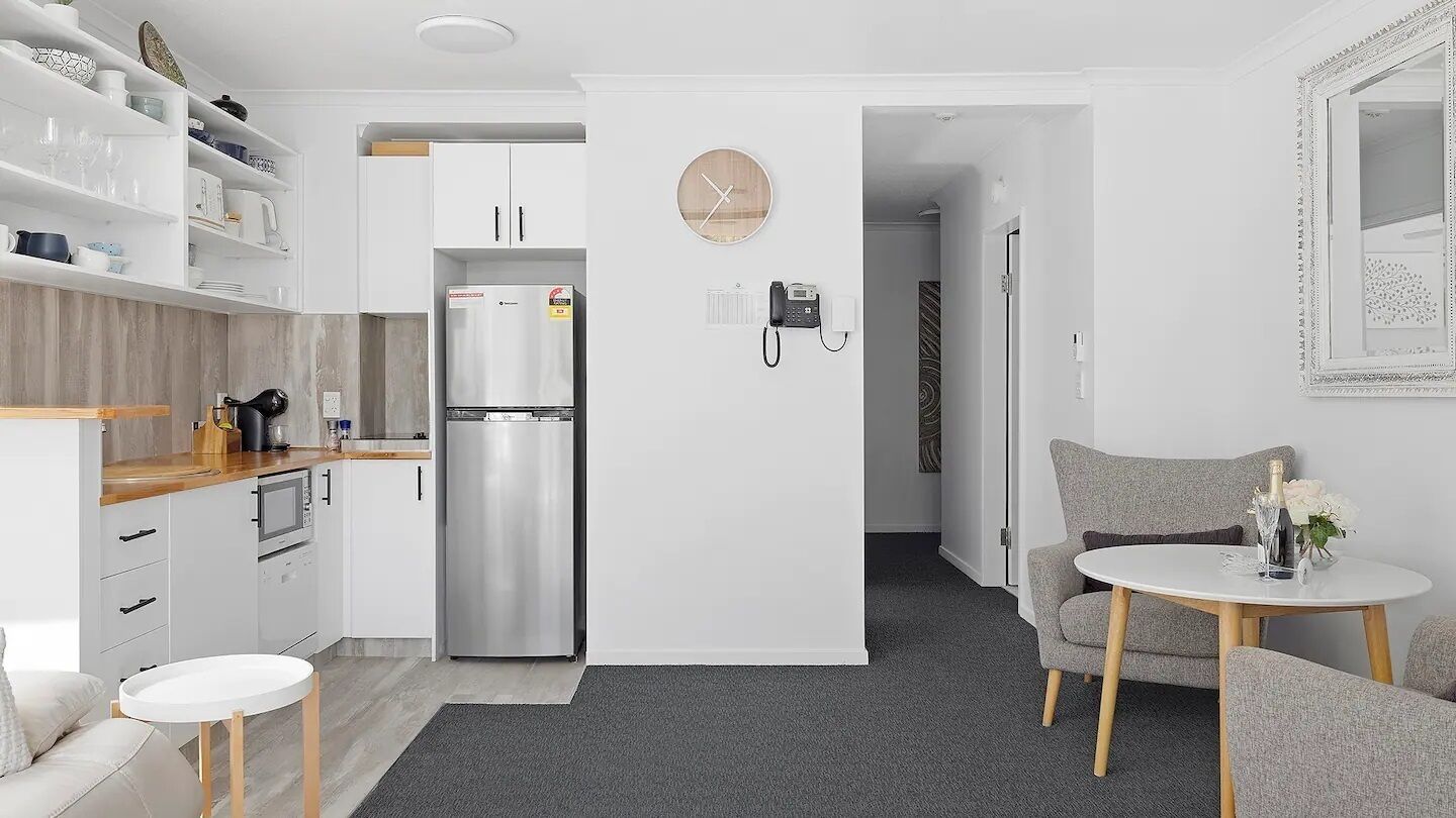 A modern kitchen with white cabinets, a stainless steel refrigerator, and a wooden countertop. There is a small round table with a chair in the foreground.