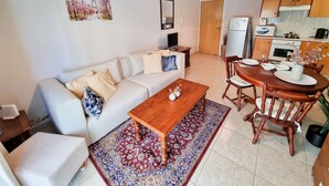 A living room with a sofa, coffee table, and dining table set with chairs. There is a kitchen area with cabinets and appliances. - Dock Holiday Pool Apartment (Larnaca)