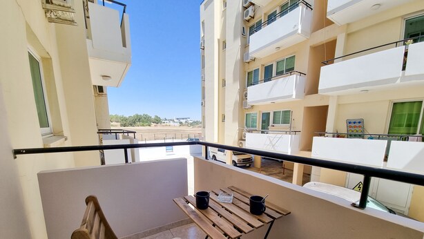 A balcony with two wooden chairs and a small table, overlooking a parking area and adjacent buildings. - Dock Holiday Pool Apartment (Larnaca)