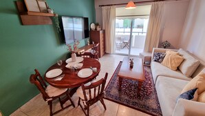 A dining area with a wooden table and chairs, a TV mounted on the wall, and a sofa in the living area. - Dock Holiday Pool Apartment (Larnaca)