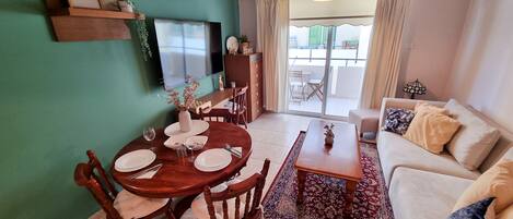 A dining area with a wooden table and chairs, a TV mounted on the wall, and a sofa in the living area.