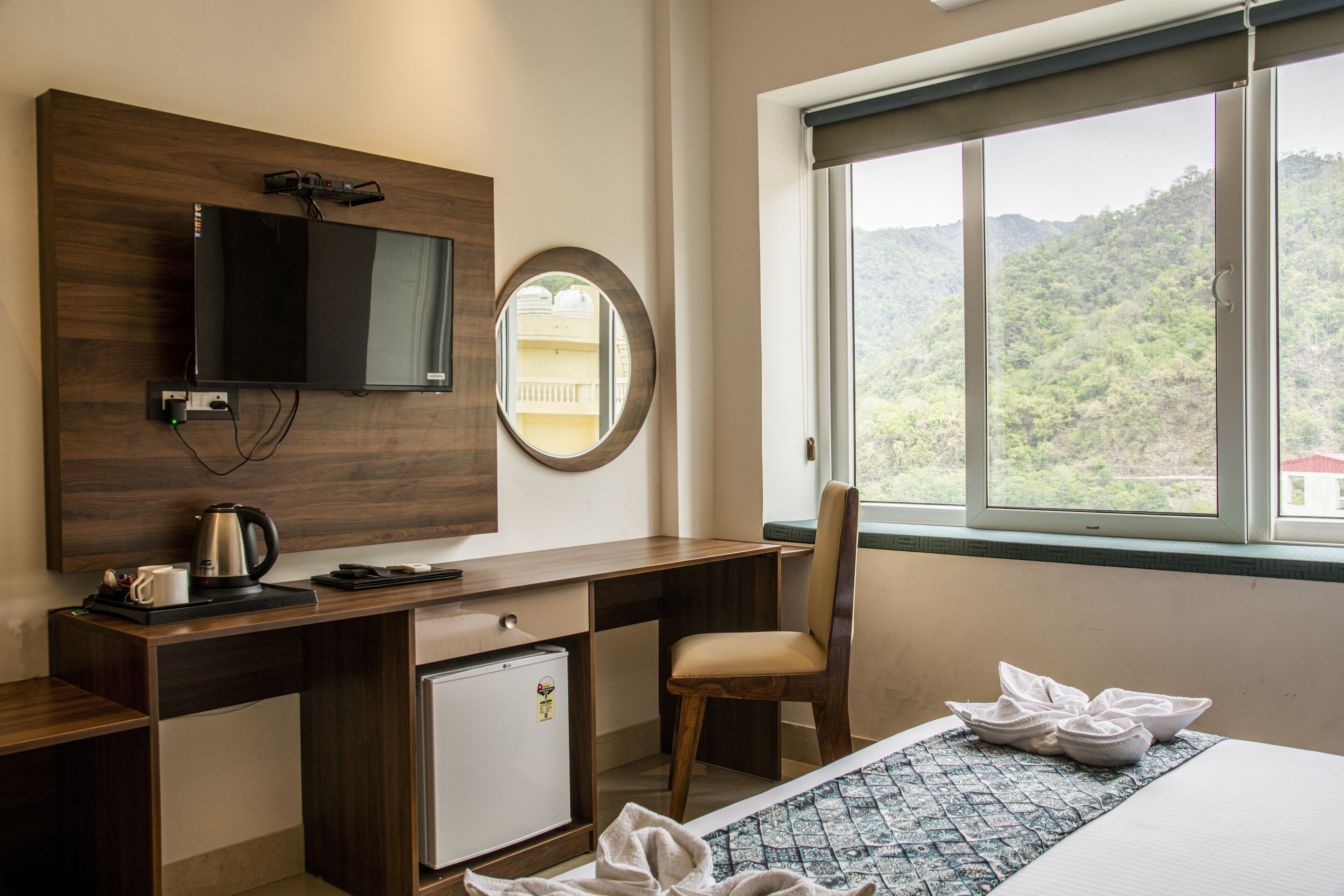 Habitación de hotel con escritorio de madera, televisor, refrigerador pequeño, silla, cama con colcha estampada en azul y blanco, y ventana con vistas a un paisaje montañoso.