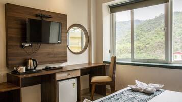 Habitación de hotel con escritorio de madera, televisor, refrigerador pequeño, silla, cama con colcha estampada en azul y blanco, y ventana con vistas a un paisaje montañoso.