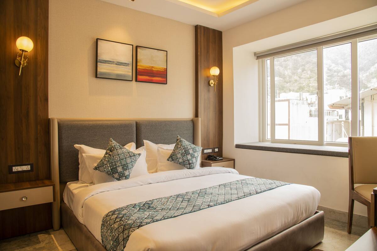 Une chambre d’hôtel avec un grand lit, deux cadres accrochés au mur, une table de chevet avec une lampe et une fenêtre donnant sur des immeubles.