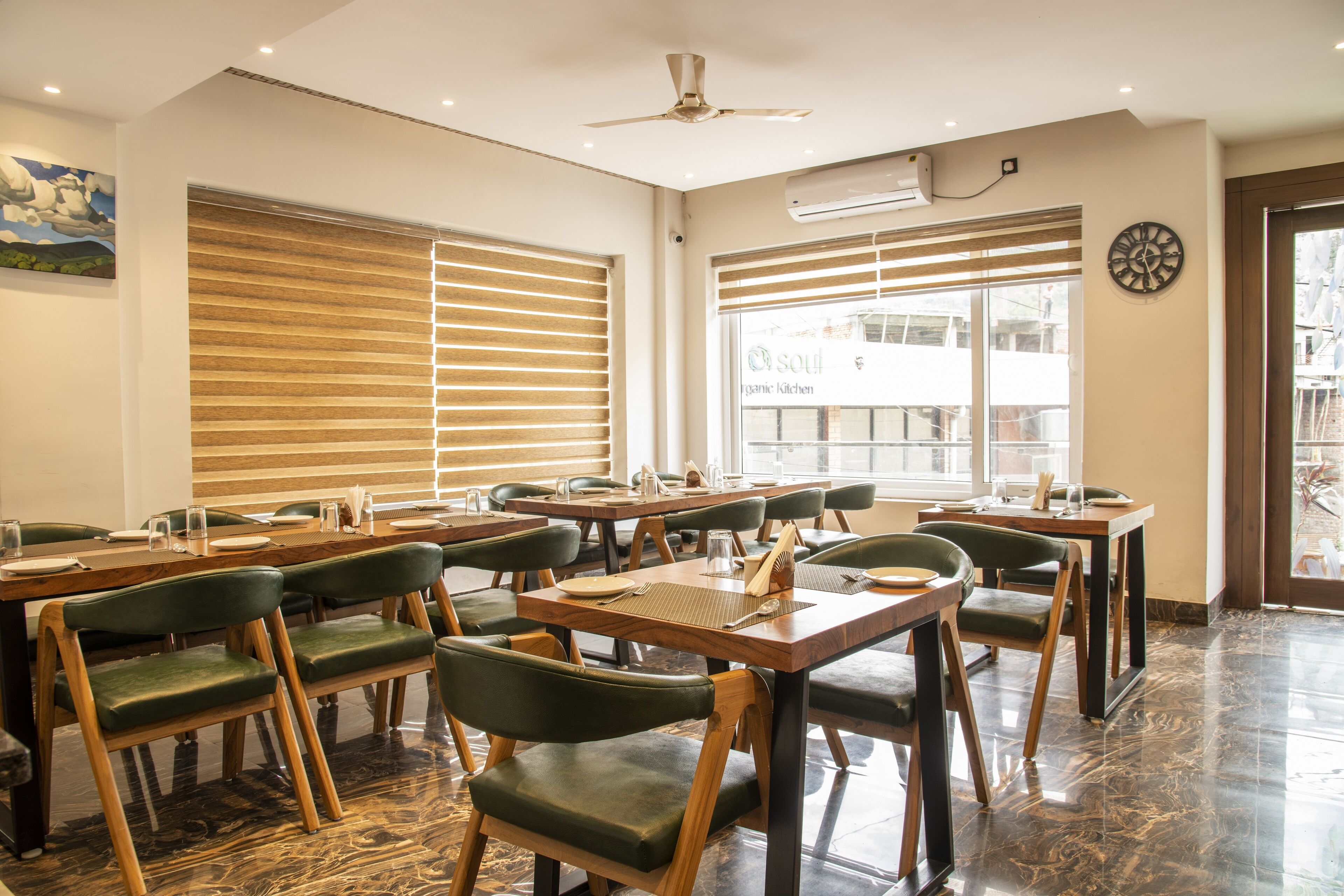 A restaurant with wooden tables and green chairs, large windows with blinds, and a ceiling fan.