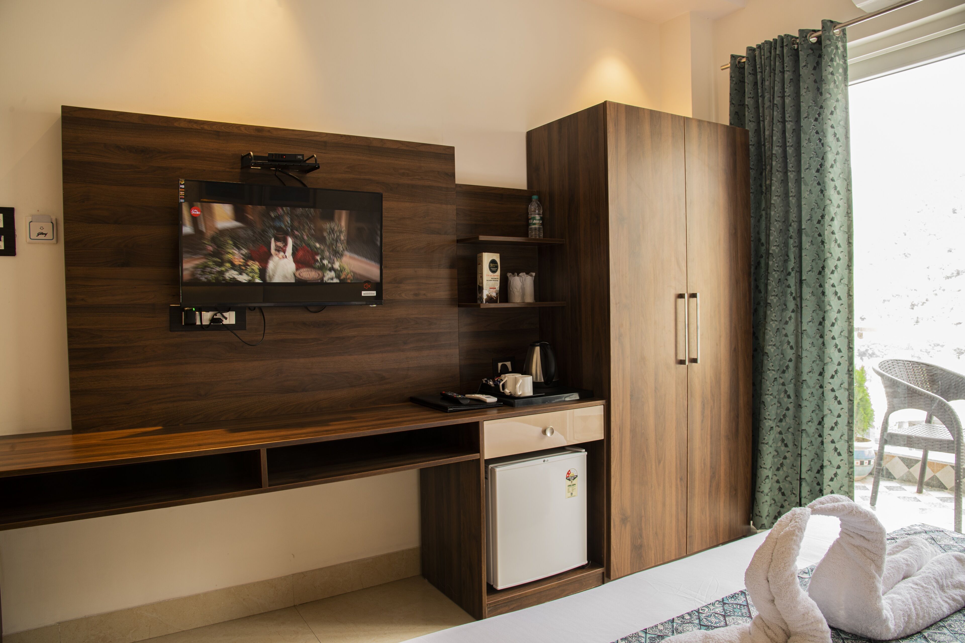 Une chambre d’hôtel moderne dotée d’un téléviseur à écran plat fixé à un mur en bois, d’un petit réfrigérateur blanc, d’une armoire en bois et d’un balcon avec un fauteuil en osier.