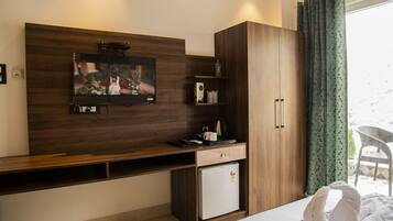 Une chambre d’hôtel moderne dotée d’un téléviseur à écran plat fixé à un mur en bois, d’un petit réfrigérateur blanc, d’une armoire en bois et d’un balcon avec un fauteuil en osier.