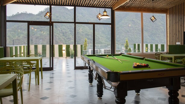A pool table in a room with large windows, wooden ceiling, and green chairs. - Hotel Dream Inn (Narendranagar)
