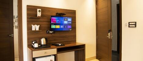 Habitación de hotel con escritorio de madera, televisor montado en la pared, refrigerador pequeño y cama con manta estampada.