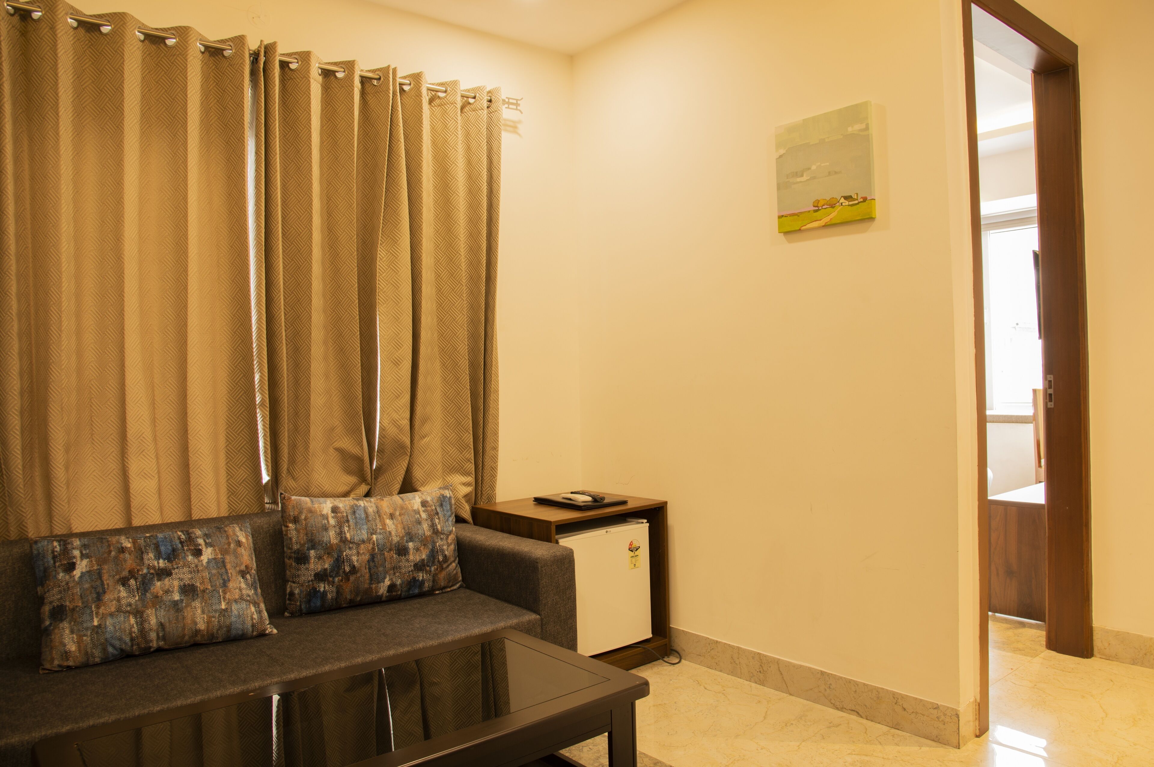 Una sala de estar con un sofá, almohadones, una mesita, una mesita auxiliar con un dispositivo y una puerta que da a otra habitación.