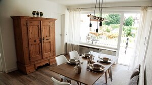 Un coin repas avec une table en bois dressée pour quatre personnes, des chaises blanches, un buffet en bois et une vue sur l’extérieur par une fenêtre ornée de rideaux légers.