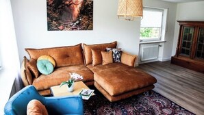 Un salon avec un canapé d’angle marron, un fauteuil bleu, un tapis à motifs, un luminaire suspendu en fibres tressées et un tableau encadré au mur.