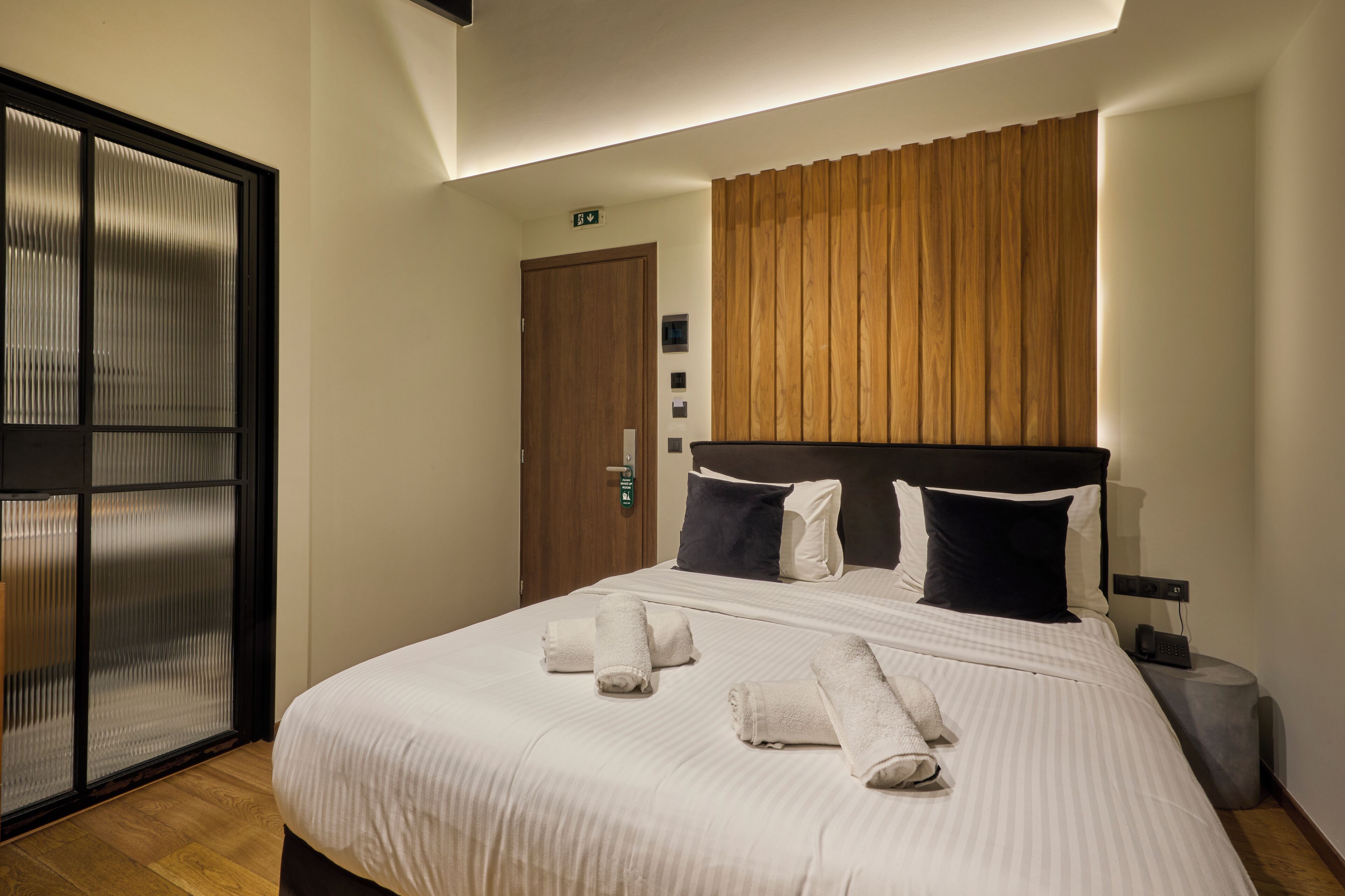 A hotel room with a large bed, a wooden headboard, a door with a handle, and a small bedside table.