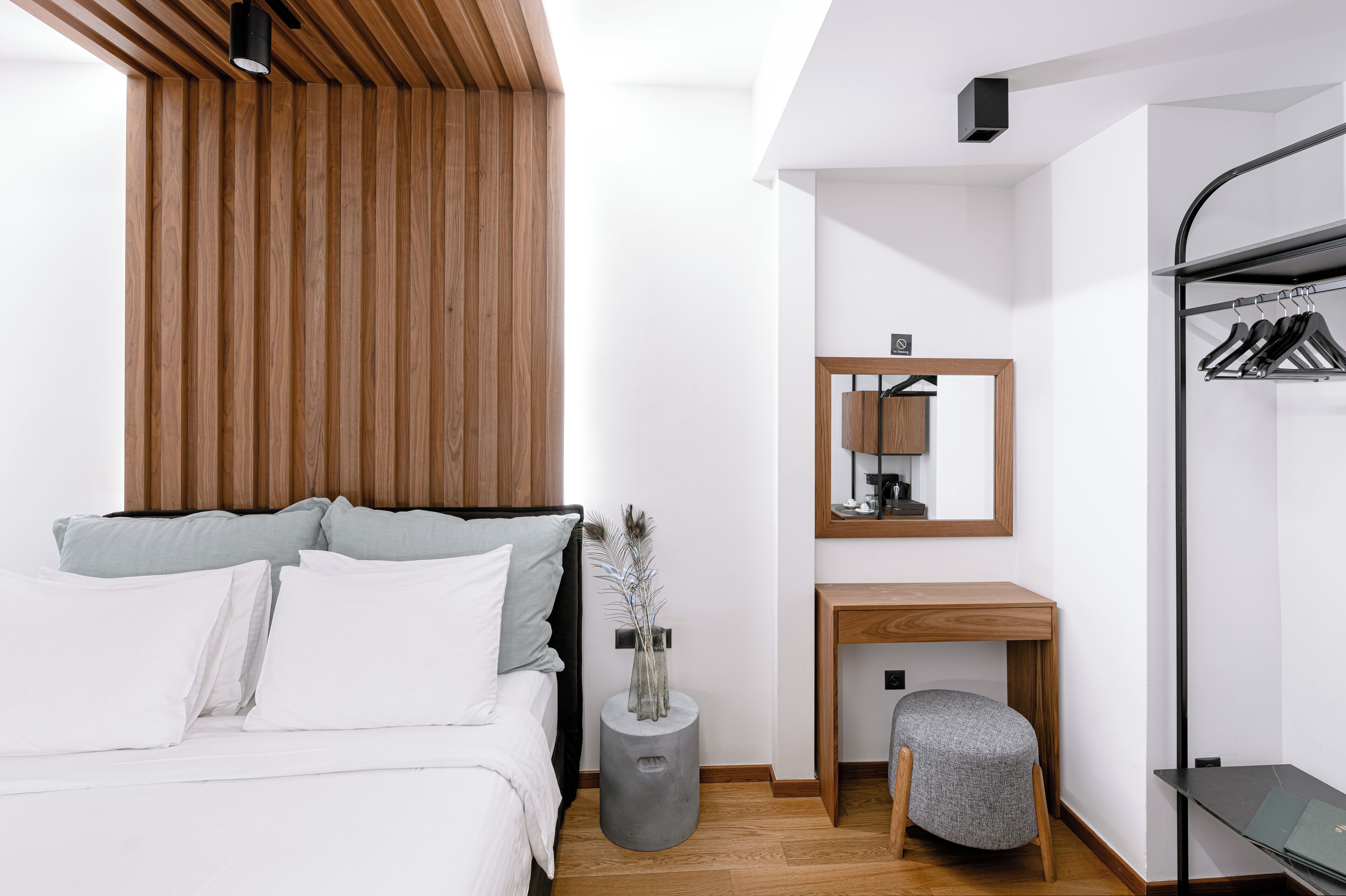 A modern bedroom with a wooden slatted headboard, a bedside table with a mirror, a grey bench, and a clothing rack.