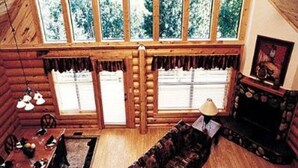A log cabin interior with a dining area, a fireplace, and large windows offering a view of a forest. - Entire Charming Cabin With Lake And Mountain View (Angel Fire)