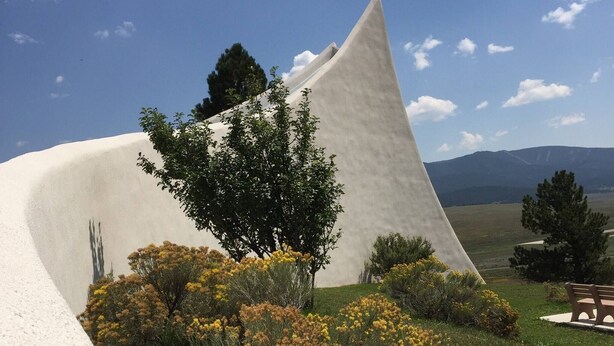 A modern building with a unique triangular design, surrounded by a grassy area with yellow flowers and a bench. - Entire Charming Cabin With Lake And Mountain View (Angel Fire)