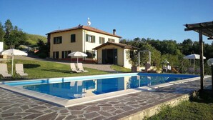 Ein Pool mit Steinplatten, ein Haus mit Ziegeldach und eine Rasenfläche mit Liegestühlen und Sonnenschirmen.