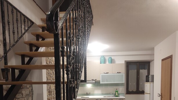 A living room with a beige sofa, a wooden staircase with black railings, a kitchen area with cabinets and a refrigerator, and a door leading to another room. - Casa Moratto in the Heart of Buje (Buje)