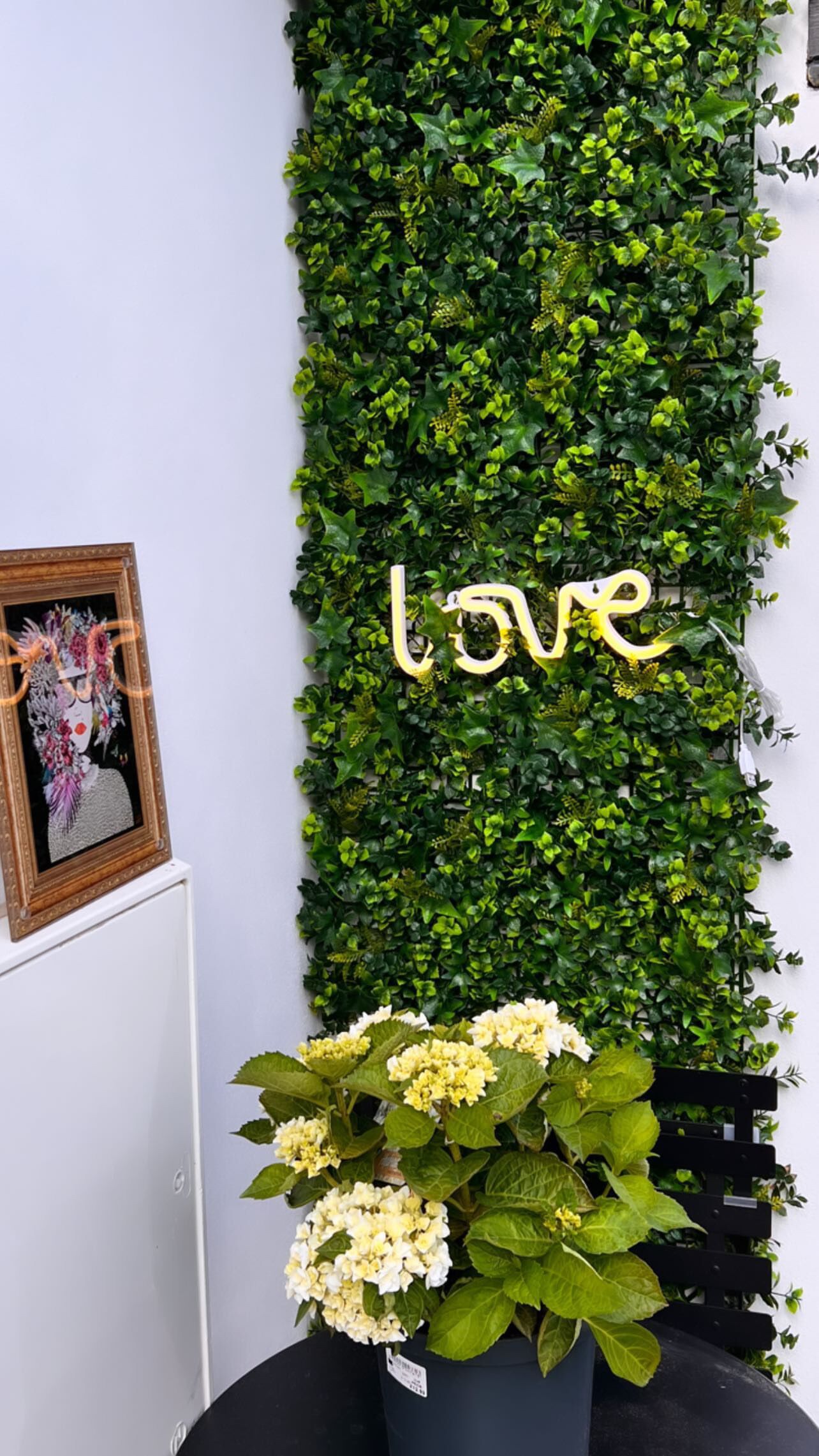 A wall with green foliage and a 'love' sign, a framed artwork, and a potted plant with white flowers.