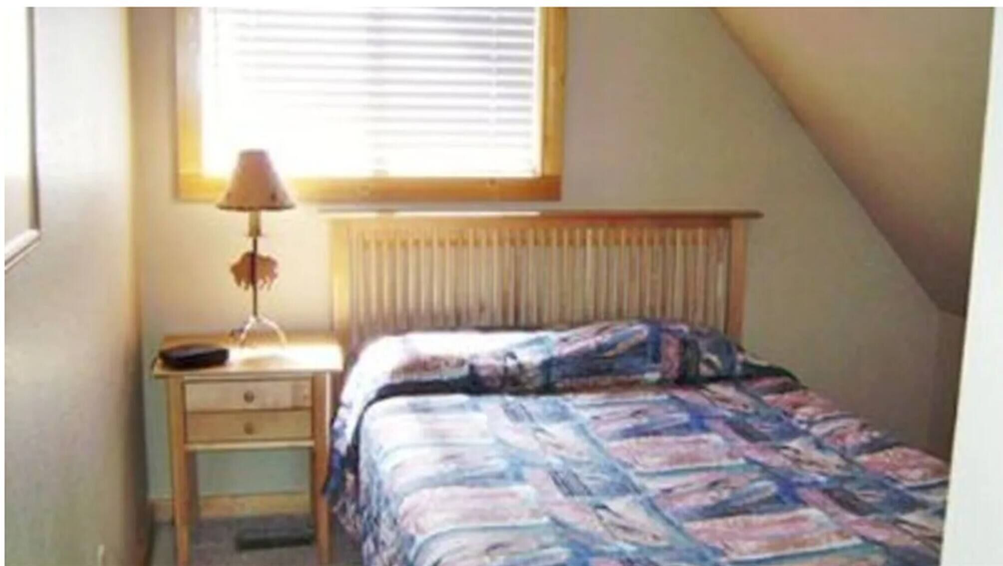 Ein kleines, schlichtes Schlafzimmer mit einem Einzelbett, einem Nachttisch, einer Lampe und einem Fenster mit Jalousien.