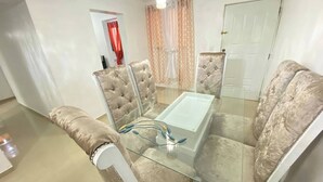 Un coin repas avec une table en verre et des chaises à capitonnage, un lustre et une porte visible au fond.