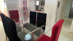 Een eetruimte met een glazen tafel, rode en zwarte stoelen met bekleding, een kroonluchter en een keuken op de achtergrond.