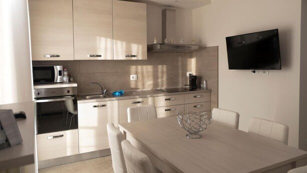 Une cuisine moderne avec des meubles en bois clair, des appareils en acier inoxydable, un téléviseur à écran plat fixé au mur et un coin repas avec une table et des chaises blanches.