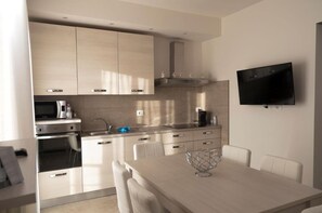 Une cuisine moderne avec des meubles en bois clair, des appareils en acier inoxydable, un téléviseur à écran plat fixé au mur et un coin repas avec une table et des chaises blanches.