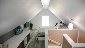 Quarto com teto inclinado, cama com cabeceira branca, mesa de cabeceira, abajur e janela.