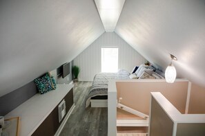 Quarto com teto inclinado, cama com cabeceira branca, mesa de cabeceira, abajur e janela.